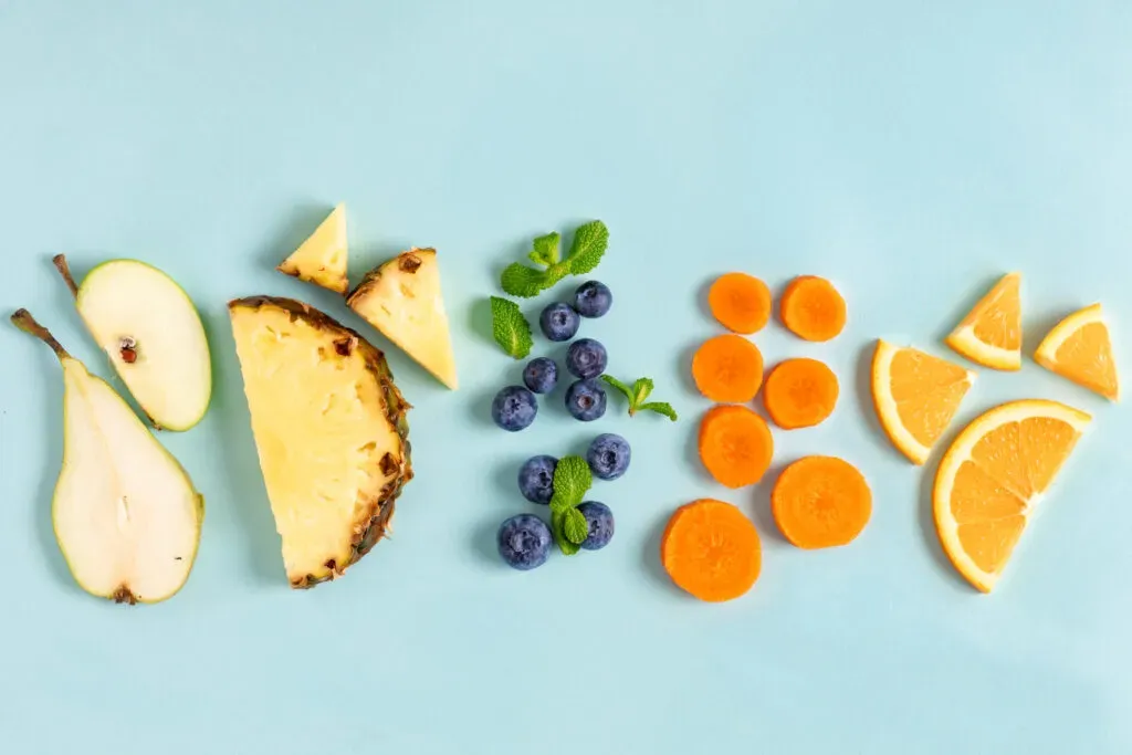 Pokrojone owoce i warzywa – gruszka, ananas, borówki, marchew i pomarańcza ułożone na błękitnym tle – ilustracja do sekcji żywność, pasze i karmy w kancelarii KRK